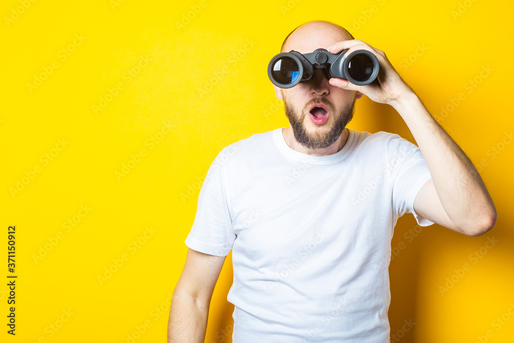 Young man with a beard looks in surprise through binoculars on a yellow background