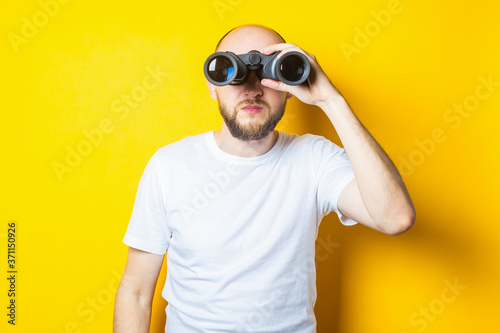Young man with a beard looking through binoculars on a yellow background © Alex