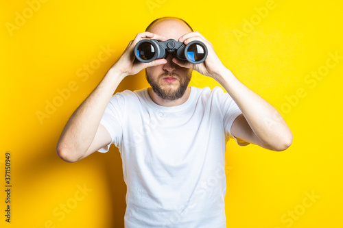 Young man with a beard looking through binoculars on a yellow background.