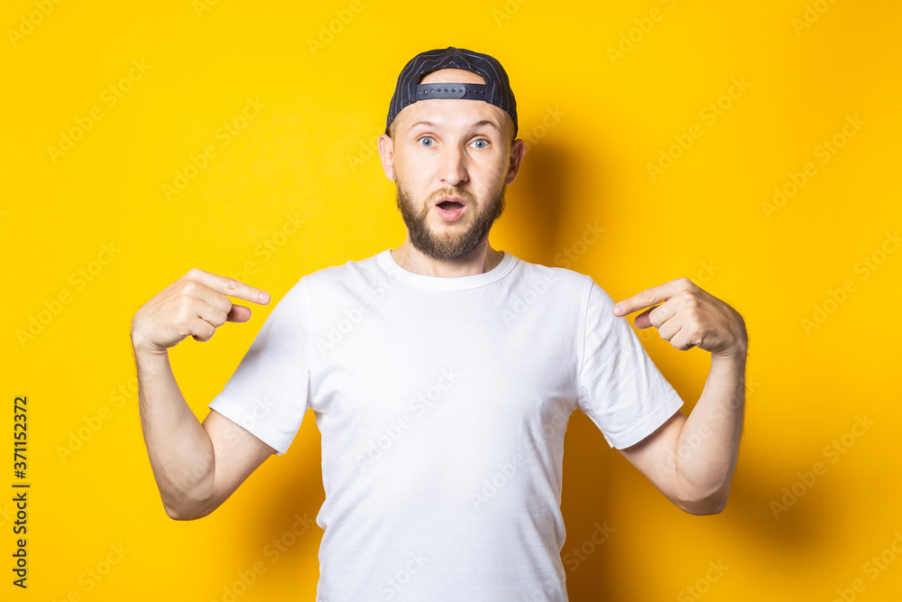 Surprised young man with beard in white clean t-shirt points fingers at himself on yellow background. Surprised young man with beard in white clean t-shirt points fingers at himself on yellow background.