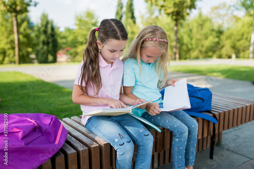 Two cute schoolgirls with backpacks and notebooks sitting on a bench near the school and reading exercises in books. Pupils studying homework together outside. Back to school concept