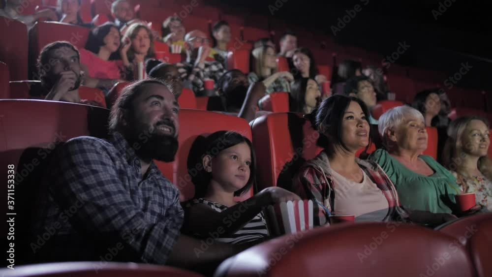Excited diverse viewers watching interesting movie in cinema. Bearded dad, teenage girl, mom and grandma chatting, eating popcorn and drinking soda without looking up from cinema screen