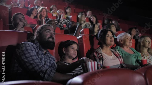 Excited diverse viewers watching interesting movie in cinema. Bearded dad, teenage girl, mom and grandma chatting, eating popcorn and drinking soda without looking up from cinema screen