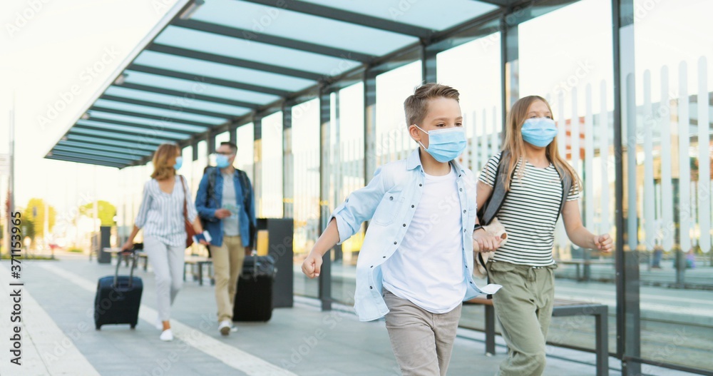 Cute small Caucasian happy kids, brother and sister in medical masks running at train station. Parents walking on blurred background and carrying suitcases on wheels. Trip concept in pandemic. Cute small Caucasian happy kids, brother and sister in medical masks running at train station. Parents walking on blurred background and carrying suitcases on wheels. Trip concept in pandemic.