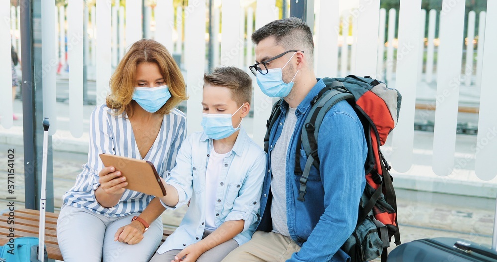 Caucasian mother and father sitting on bench at bus stop with small son and using tablet device. Parents with kid in medical masks waiting for transpost with suitcases and looking for route on gadget.