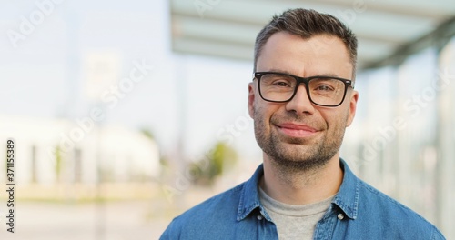 Portrait of handsome Caucasian man in glasses looking straight and smiling to camera outdoor. Atractive happy male in eyeglasses at bus stop on summer sunny day. Close up. Zooming in. © VAKSMANV