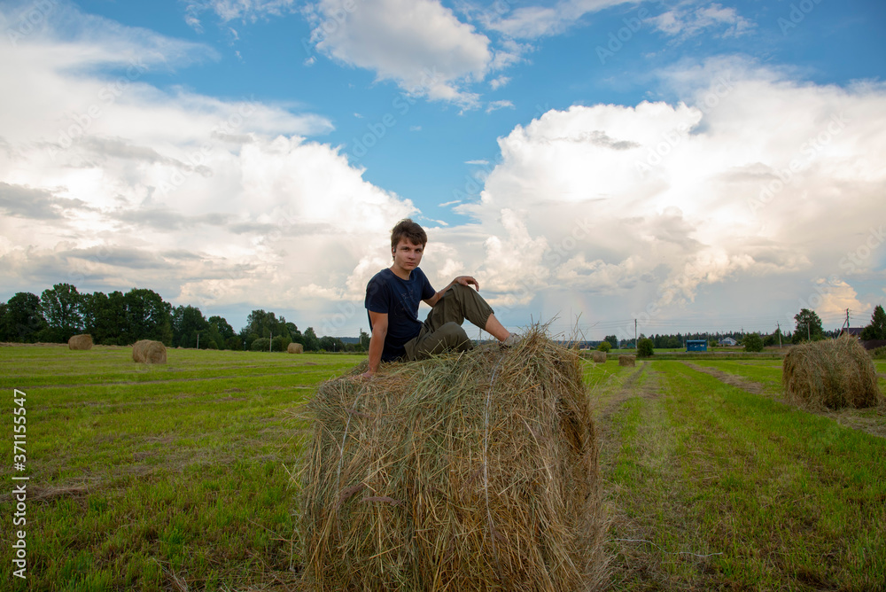 A handsome young man is sitting on a haystack against a blue cloudy sky. A handsome young man is sitting on a haystack against a blue cloudy sky.