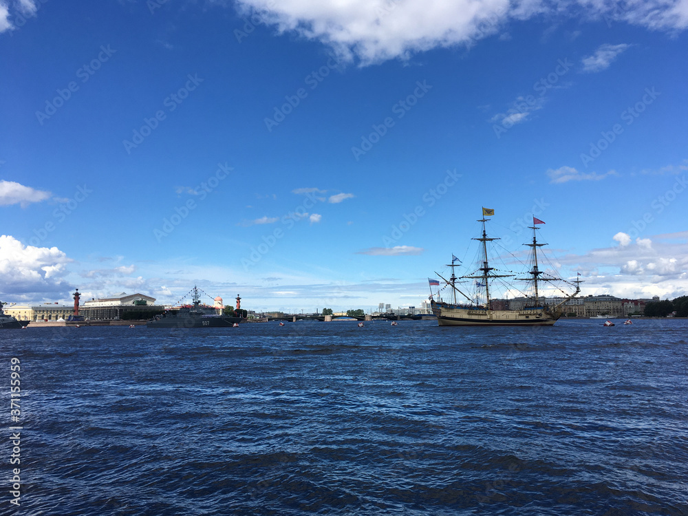The Neva River with the sailing ship Poltava and warships lined up for the naval parade in St. Petersburg against the background of a blue sky with clouds.