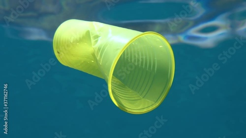 Slow motion. Plastic pollution, yellow plastic cup drifting under surface of the blue water in sun lights. Plastic garbage environmental pollution problem in seas and ocean. Adriatic Sea 