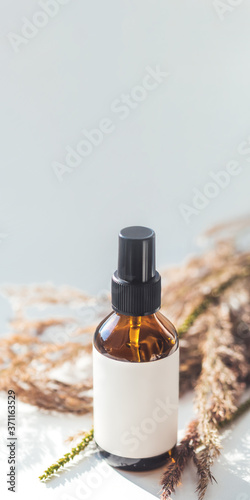 Concept of apothecary cosmetic. Herbs and brown glass bottle of essential oil in still life composition on light blue and white background. Sunlight and shadow. Natural cosmetic remedy with white tag.