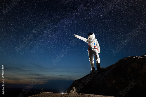 Full length of cosmonaut standing on top of rocky hill and pointing at stars. Space traveler wearing white space suit while looking at fantastic night sky. Concept of galaxy, human in space, milky way