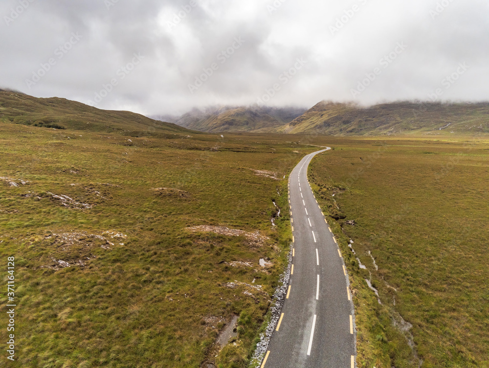 Small asphalt road into mountains. Aerial drone view, Connemara region, West of Ireland. Cloudy sky. Empty wild fields on each side of the road. Country side.