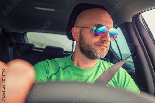 Bald man with beard driving a car looking out into window, Hands on the wheel out of focus. Driver wearing green t shirt and big reflective glasses. Concept travel, road trip.