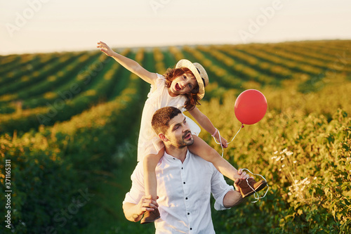 Father with his daughter that sits on shoulders and holds red balloon spending free time on the field at sunny day time of summer © standret