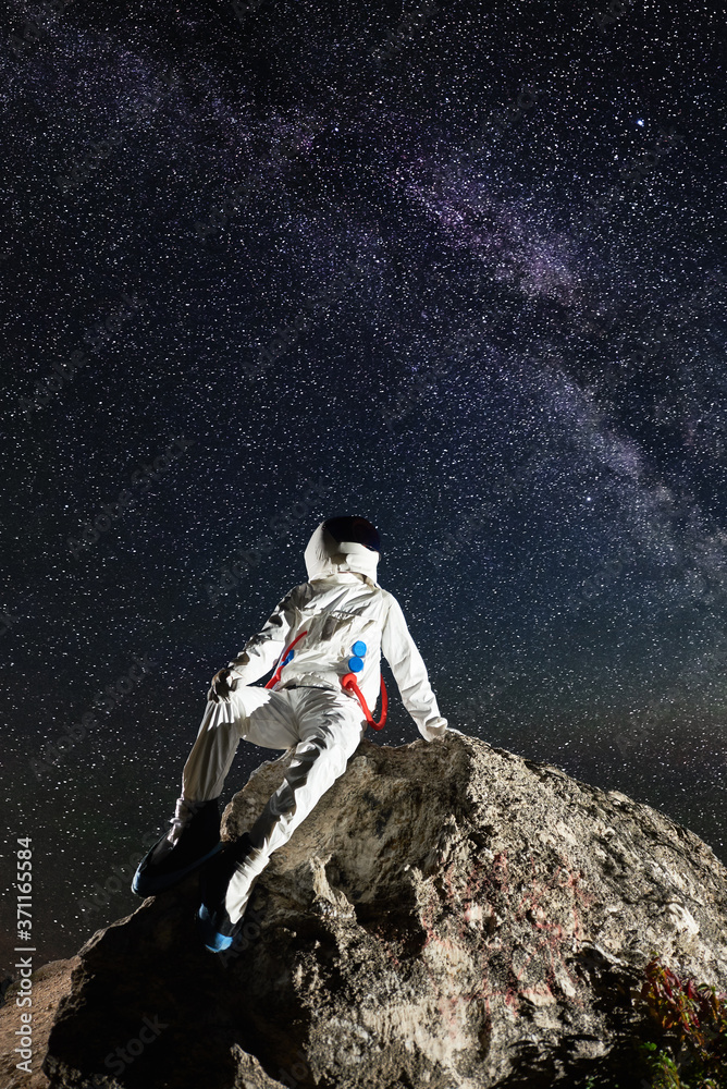 Obraz premium Astronaut resting on top of rocky mountain under beautiful night sky with stars. Astronaut wearing white space suit with helmet. Concept of space exploration, cosmonautics and milky way.