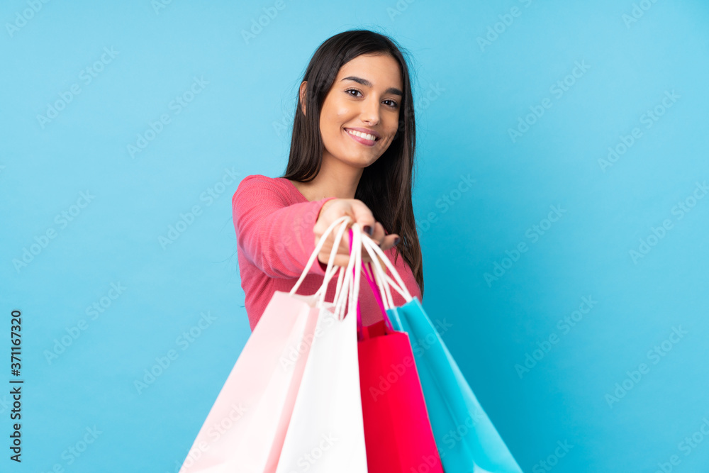 Young brunette woman over isolated blue background holding shopping bags and giving them to someone
