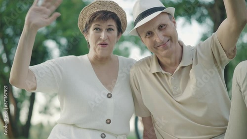 Portrait of cheerful mid-adult couple waving and smiling at camera. Happy Caucasian man and woman posing in summer park on sunny day. Joyful family enjoying weekends outdoors.