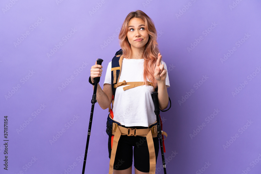 Teenager girl with backpack and trekking poles over isolated purple background with fingers crossing and wishing the best