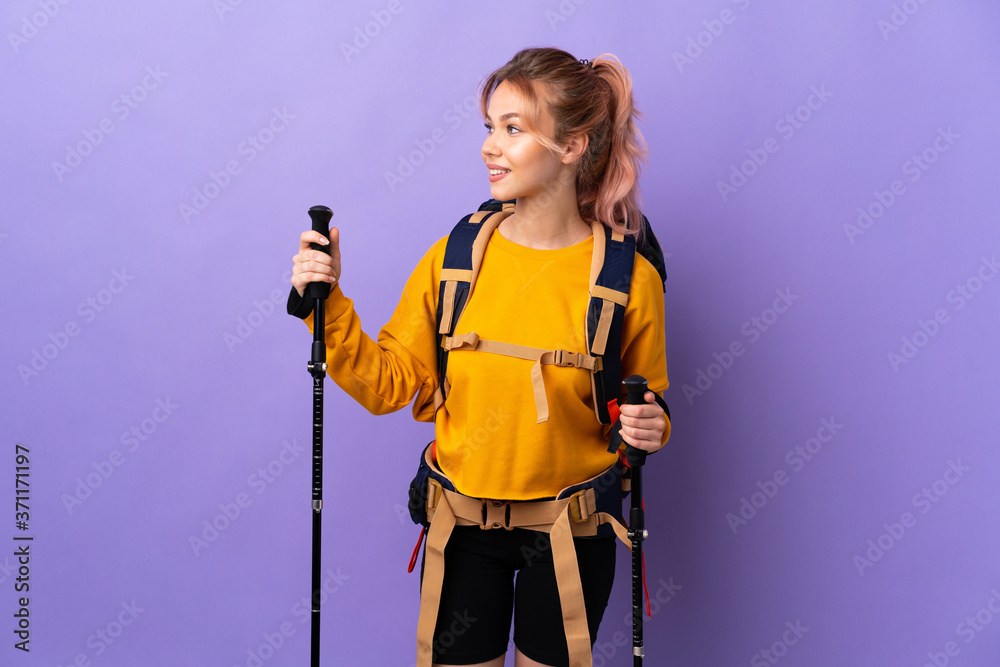 Teenager girl with backpack and trekking poles over isolated purple background looking side