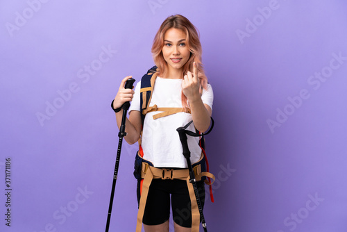Teenager girl with backpack and trekking poles over isolated purple background doing coming gesture