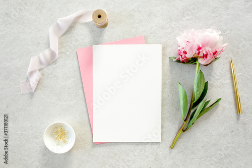 Mockup invitation, blank paper greeting card, pink envelope and peonies on gray stone table. Flower background. Flat lay, top view.