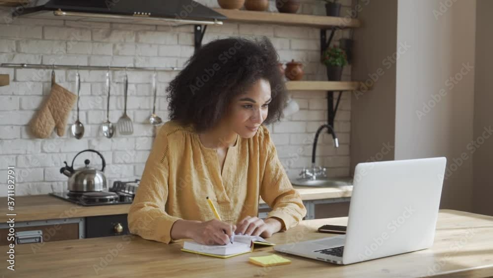 Young girl is using her laptop to study online, she is making notes while sitting on the kitchen at the table. She has an afro style hair, light cozy kitchen