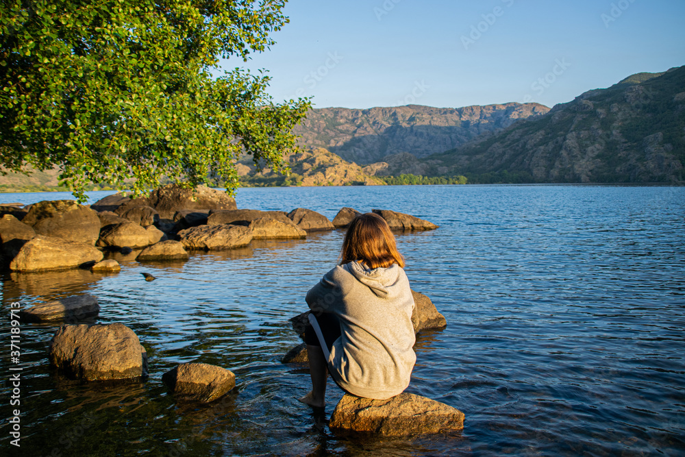 Photo of a young and attractive girl with a sweatshirt sitting on a rock natural lake during sunrise. Alone, meditation