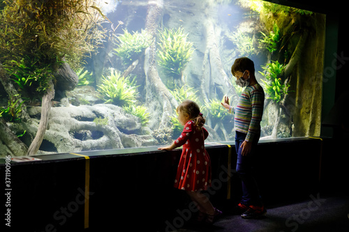 Kid boy and toddler girl visiting together zoo aquarium. Two children watching fishes, corals and jellyfishes. School child wearing medicals masks due pandemic corona virus time. Family on staycation