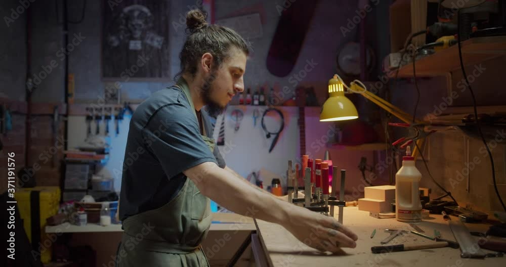 Side shot of handsome Hispanic guy happy enjoying making guitar at cozy workshop indoors, young artisan luther clamps new instrument art craftsman concept hobby of male in apron working in studio Side shot of handsome Hispanic guy happy enjoying making guitar at cozy workshop indoors, young artisan luther clamps new instrument art craftsman concept hobby of male in apron working in studio