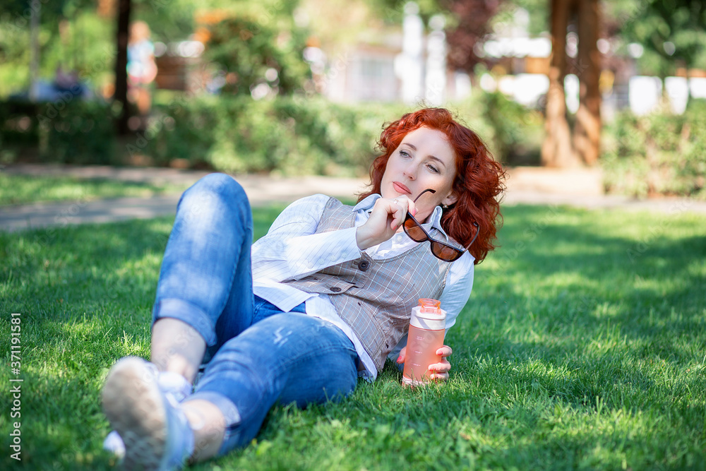 Fototapeta premium A cute red-haired girl lies in the park on the grass and thinks or dreams about something.