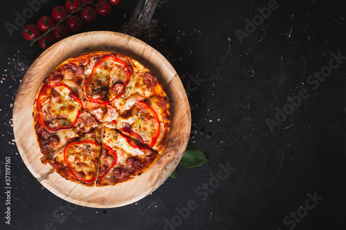 Overhead shot of Italian pizza on the wooden plate with bazil, tomatoes and salt scattered on the black background with copy space