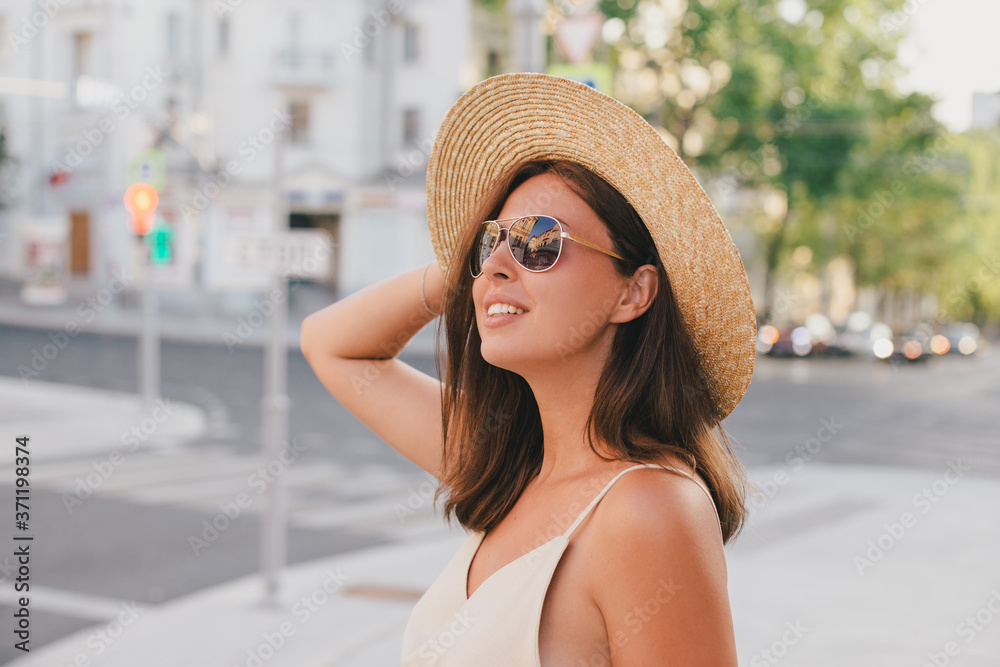 Young beautiful woman wearing straw hat, sunglasses and linen beige suit posing on a city street in old town.