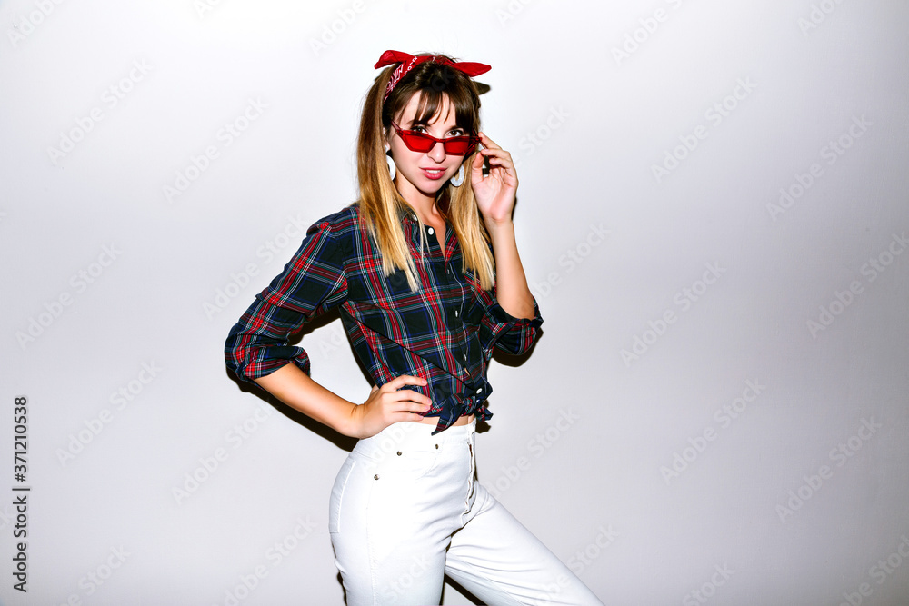 Positive indoor flash portrait of playful cheeky hipster woman showing tongue and having fun, wearing American style plaid shirt and vintage red sunglasses, trendy accessories, white background . Positive indoor flash portrait of playful cheeky hipster woman showing tongue and having fun, wearing American style plaid shirt and vintage red sunglasses, trendy accessories, white background .