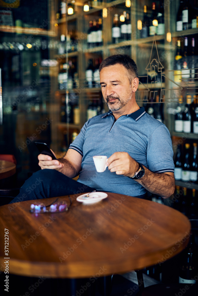 Mature man sitting in coffee shop, drinking coffee and using smart phone.