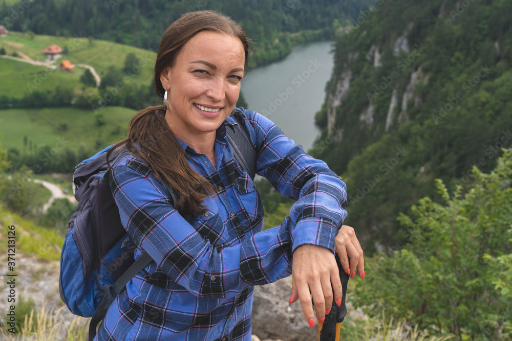 Young happy smiling woman in blue shirt, backpacker tourist, standing alone mountain ridge cliff edge, looking camera, portrait, scenic landscape view