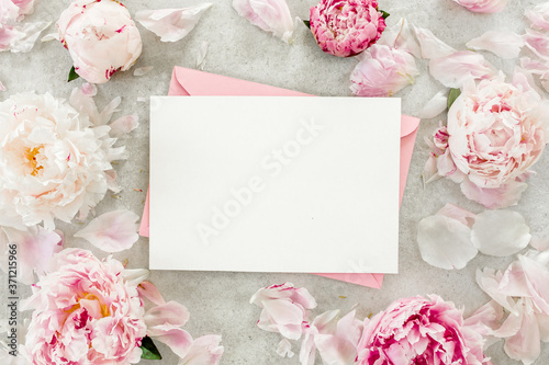 Mockup invitation, blank paper greeting card, pink envelope and peonies on gray stone table. Flower background. Flat lay, top view.