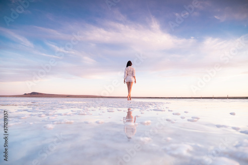 Photo of a slender woman from the back with reflection in the water. A girl walks on a salt lake at sunset.