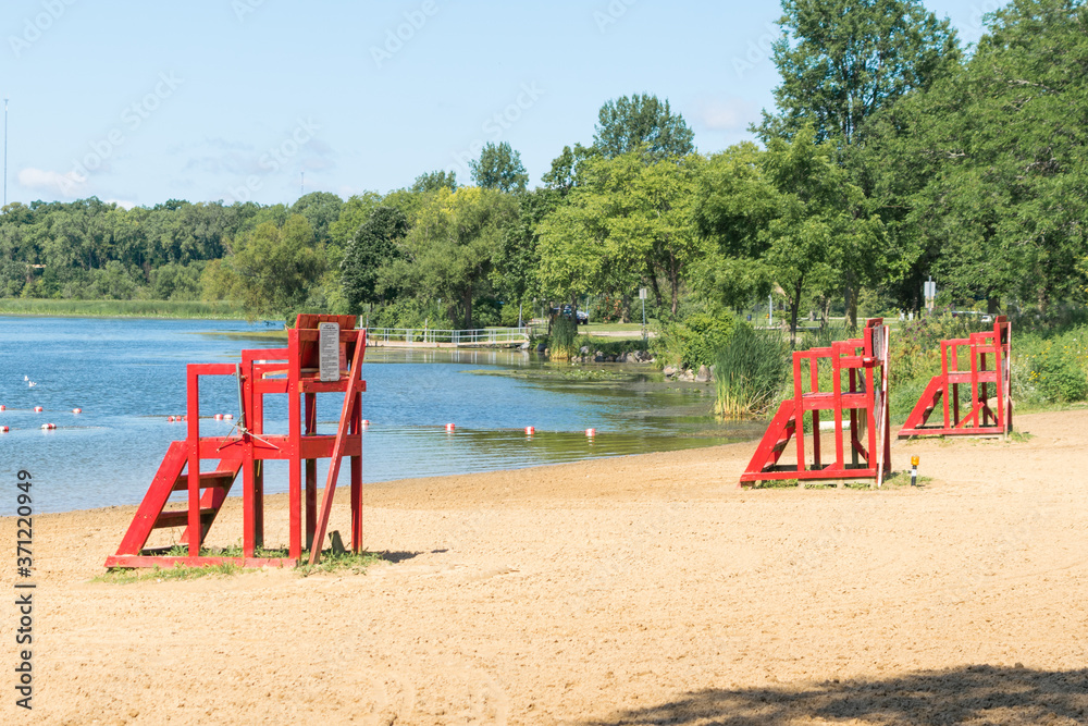 Empty beach with empty red lifeguard stands.  Clear blue lake water and sand, with no people present.  Vacant beach with empty swimming area, trees around banks.