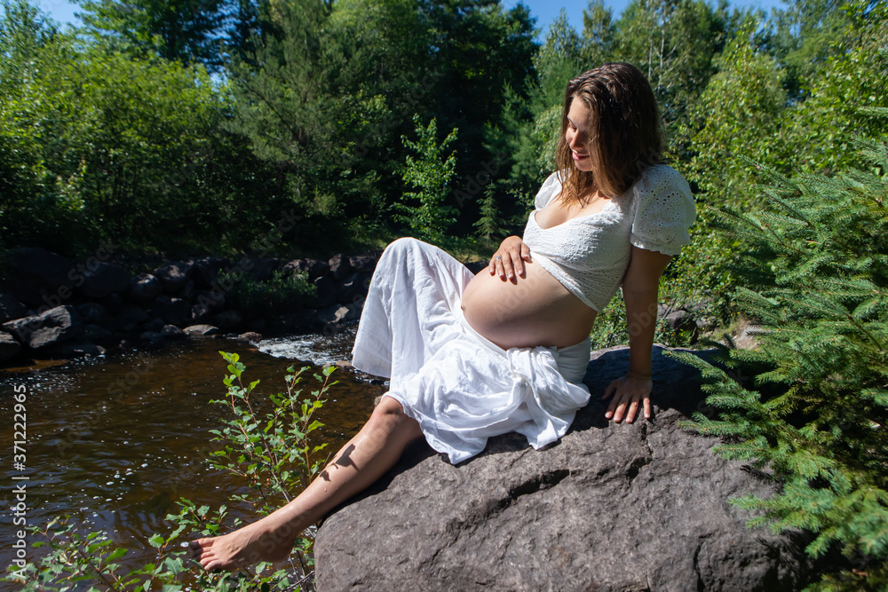 Sweet, beautiful, pregnant young woman with long hair dressed in white, sitting on a big rock next to a river and caressing her belly. Sweet, beautiful, pregnant young woman with long hair dressed in white, sitting on a big rock next to a river and caressing her belly.