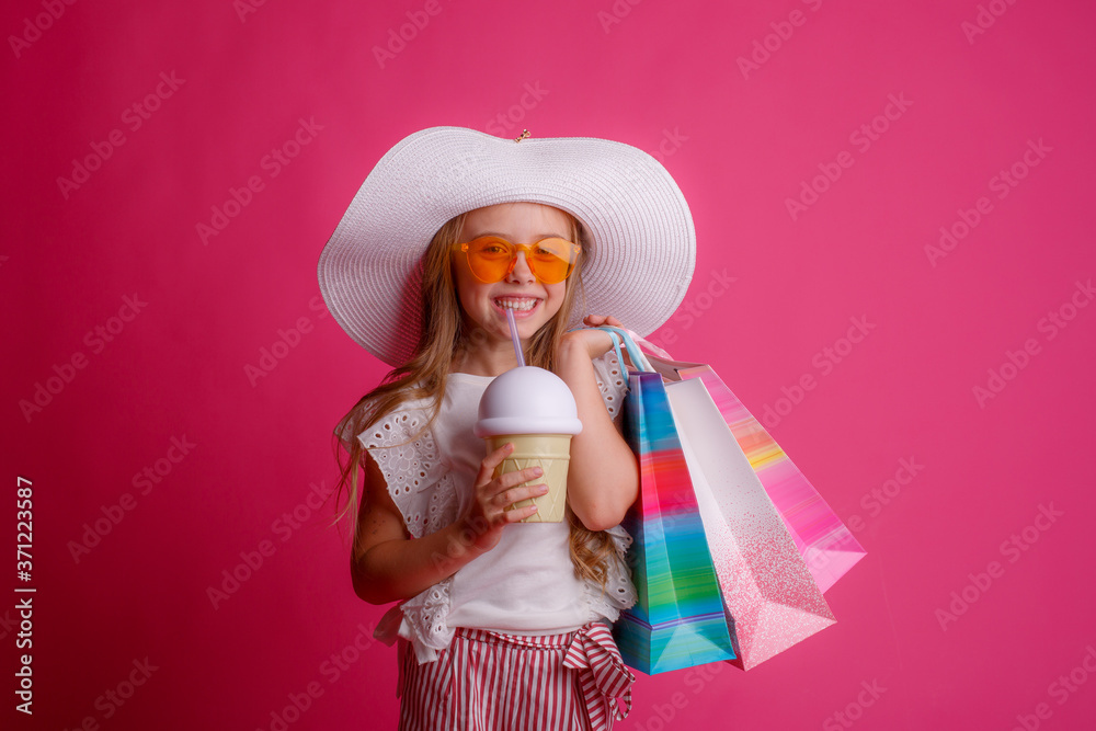 a little Girl with a lot of shopping bags, wearing sunglasses and a hat smiles on a pink background in the Studio, shopping concept, sale