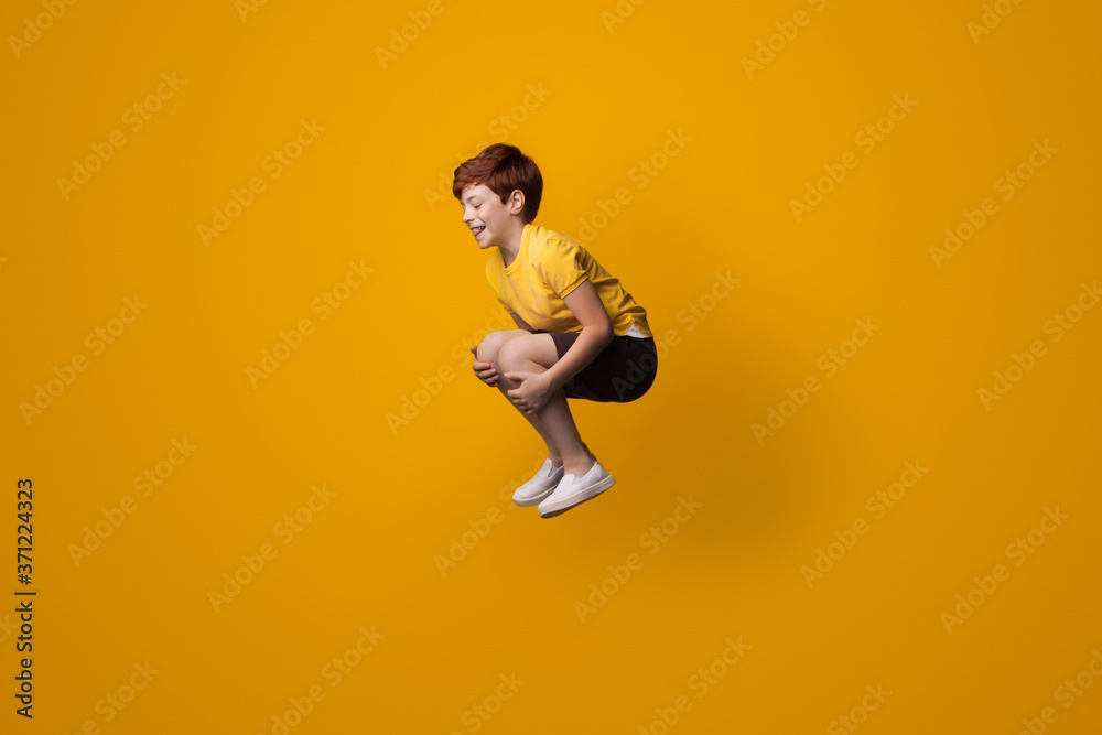 Small caucasian boy with red hair smiling and jumping on a yellow studio wall