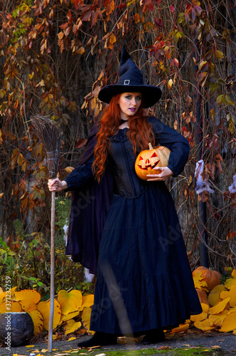 a girl in a black dress and a witch hat for Halloween with a pumpkin and a broom on a background of yellow leaves in a Park in the afternoon