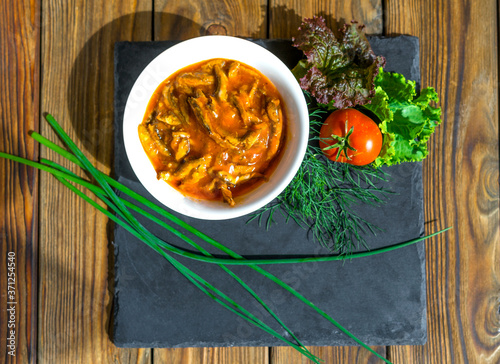 Fish in tomato sauce in a white ceramic bowl stands on a black slate Board on an old wooden table. Fish served with green onions, dill, lettuce and tomatoes. Snack, food concept. View from above. photo