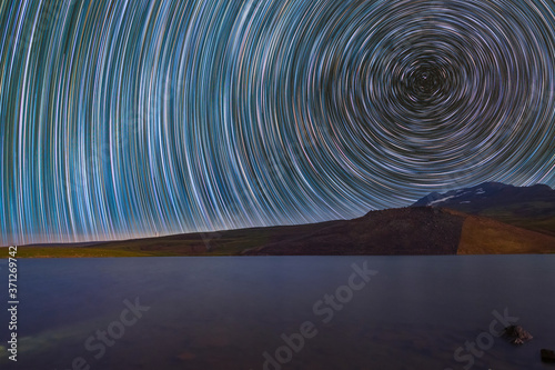 Long exposure night landpscpe. Beautiful small lake and mountains. The colorful star trails on the sky. Night time lapse photography.