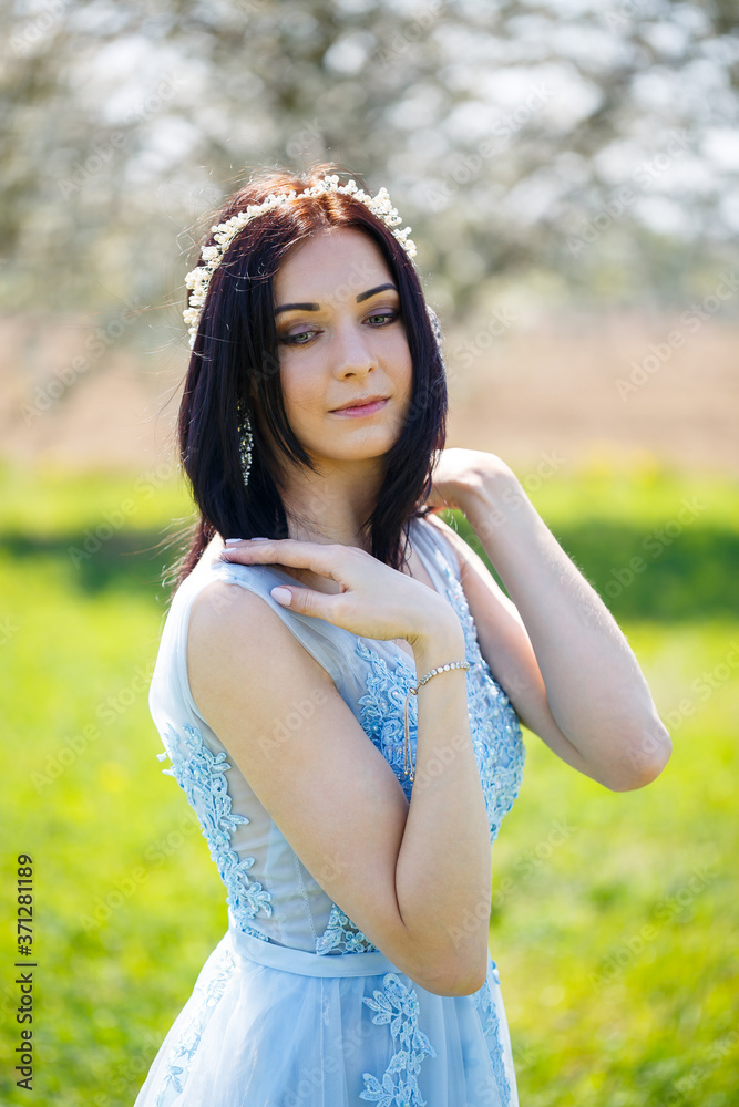 Naklejka premium Portrait of a girl in a light summer blue long dress adorned in her hair against a blossoming tree.