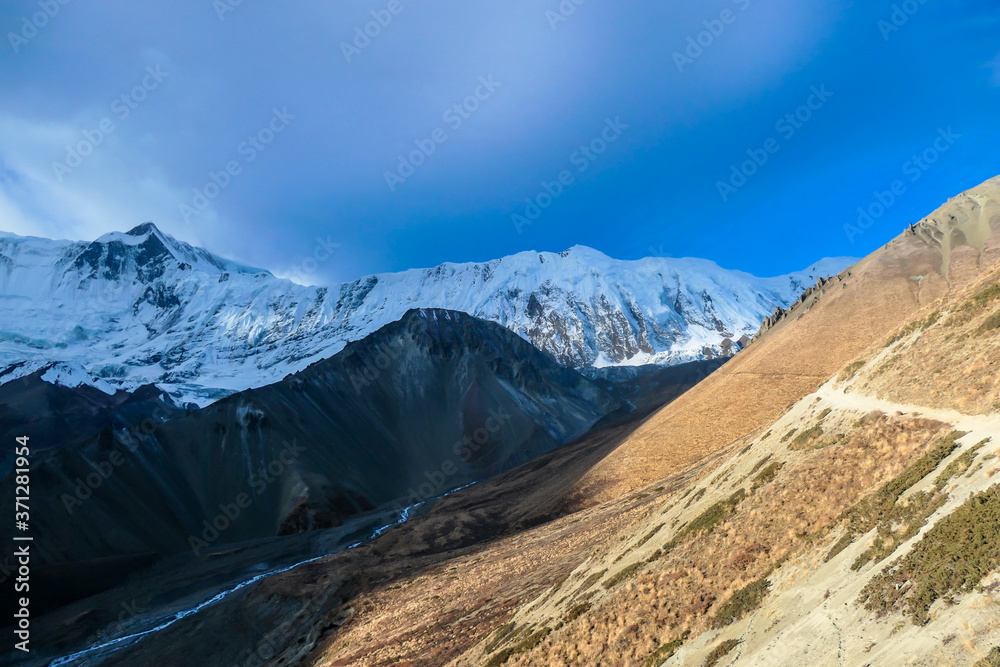 A view on the sunny pathway leading to dark, shadowy mountain peaks. Snow capped Himalayan peaks along Annapurna Circuit in Nepal, leading to Tilicho Lake. Barren and sharp slopes. New day beginning