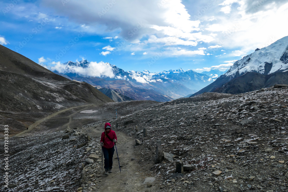 Fototapeta premium A woman in pink jacket walking on a pathway leading to Tilicho Lake, Nepal. Stormy weather in snow capped Himalayan peaks along Annapurna Circuit. Barren and sharp slopes. Coldness