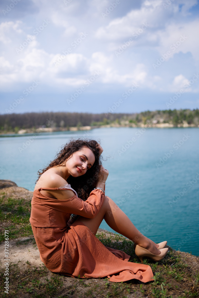A beautiful girl of European appearance with curly hair and a smile on her face sits in a green meadow against the background of a blue lake. Warm summer day, happy young woman, emotions of joy