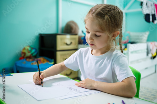 A girl in a white T-shirt sits in her room at the table and draws with colored pencils