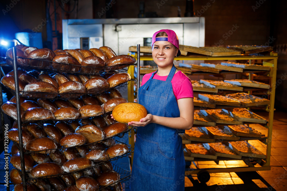 Professional baker - a young, pretty woman in a jeans apron holds fresh bread against the background of a bakery or bakery. Bakery products. Bread production
